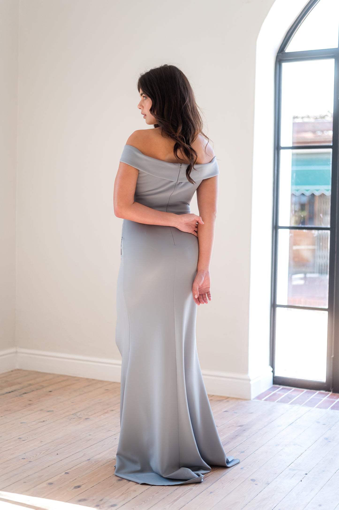 Woman in a Sage off-shoulder bridesmaid dress from the back in a light filled room.
