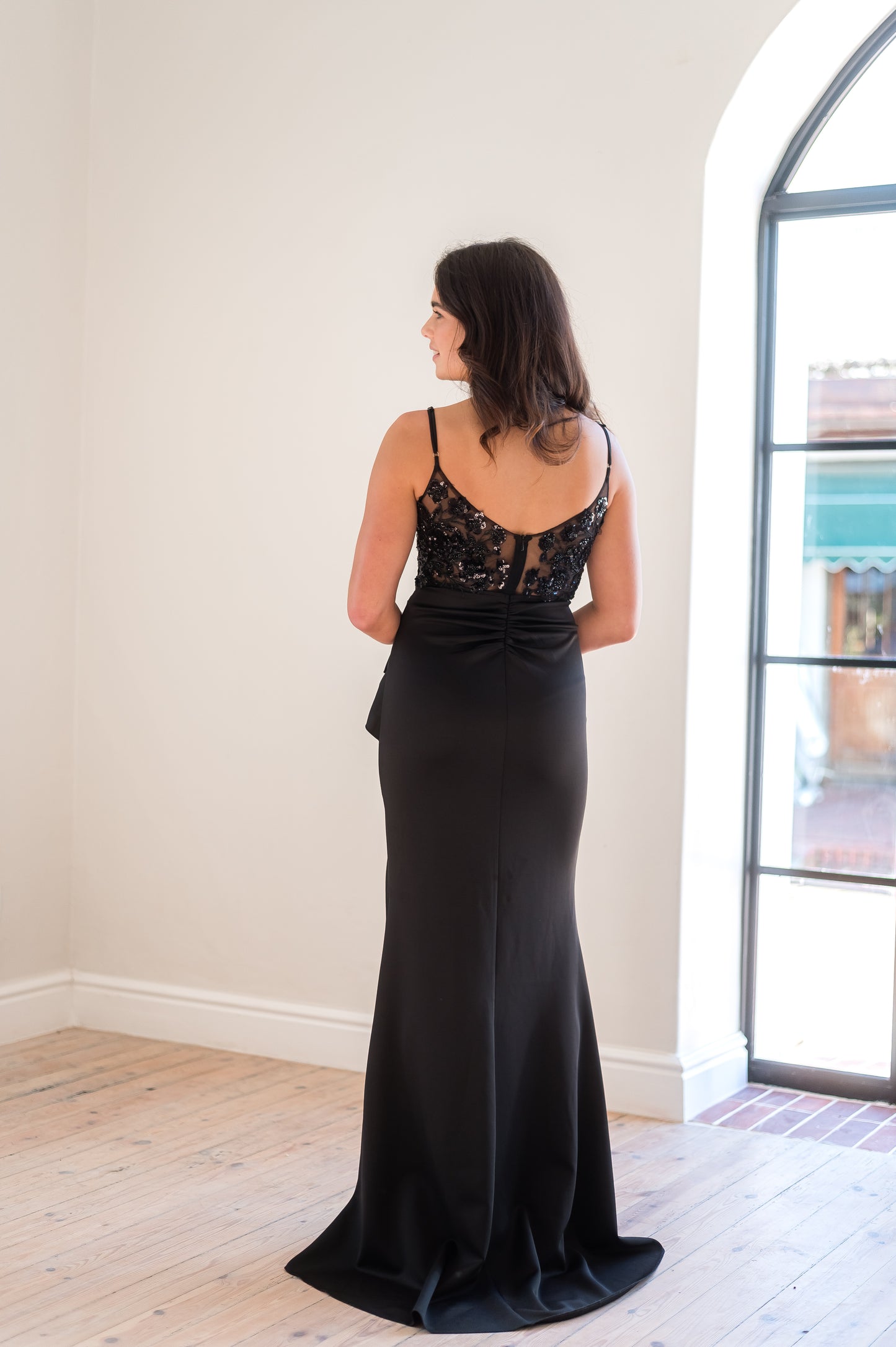 Woman wearing a black evening gown with lace detailing on the back a room with a large window.