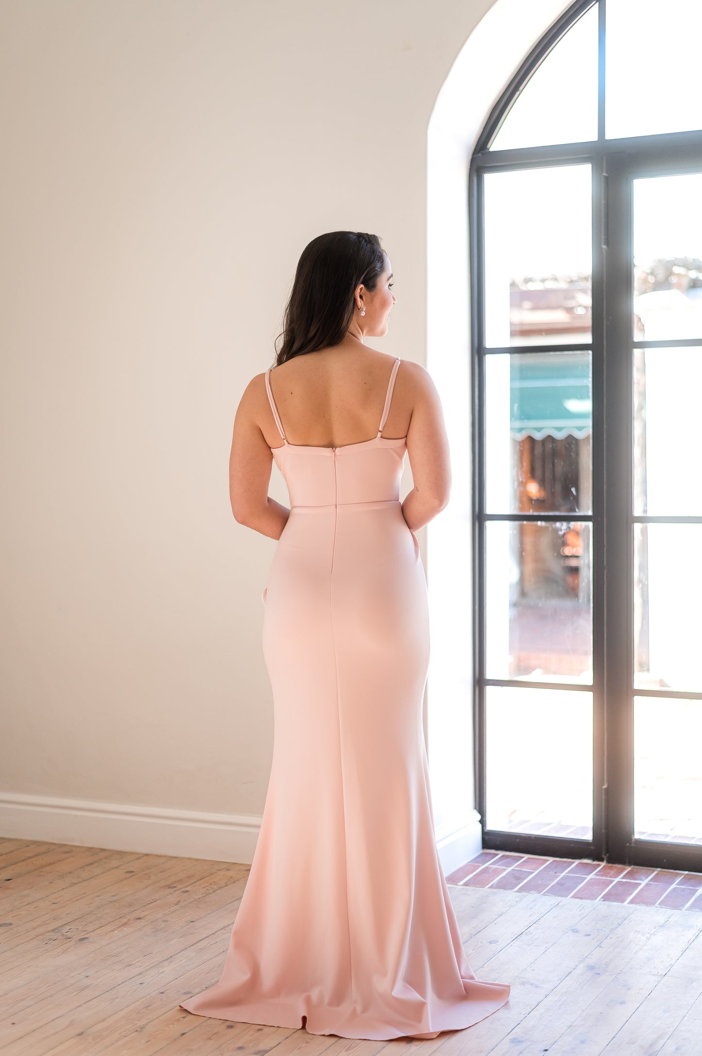 Back of a woman standing in a dusty pink , full-length bridesmaid dress with thin straps.