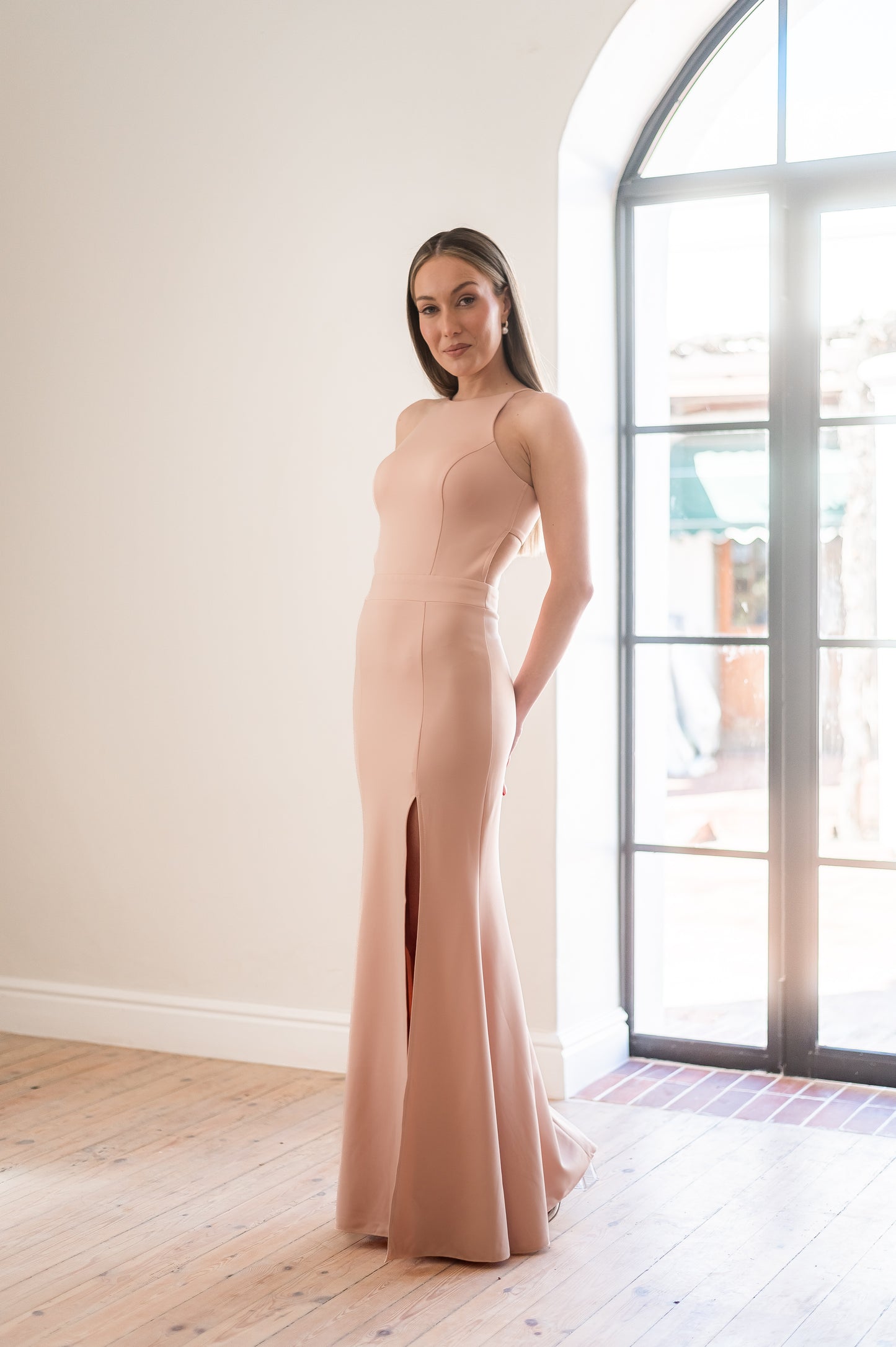 Woman in a nude halter neck bridesmaid dress with a thigh slit standing in a room with a large window.