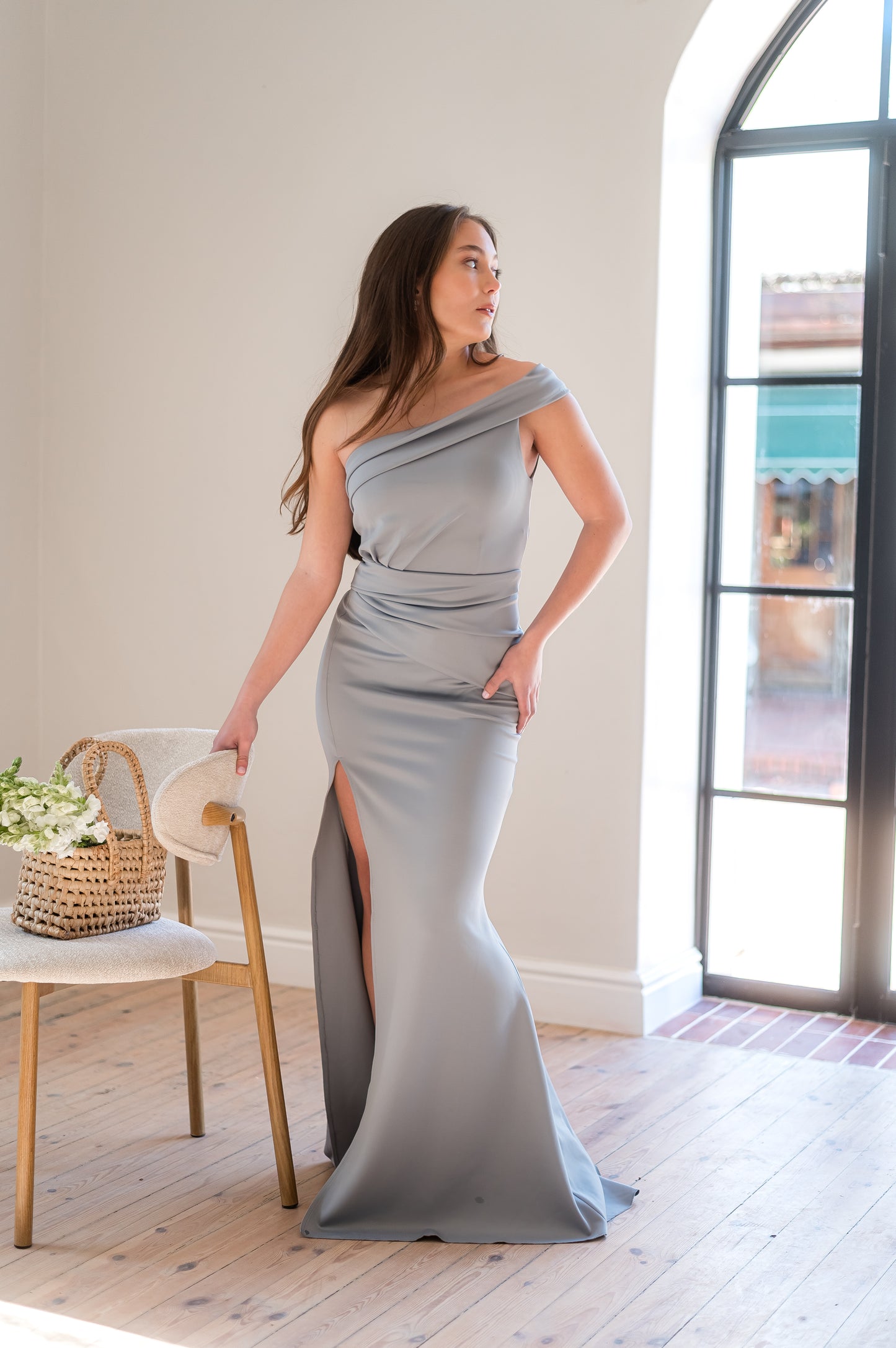 Woman in a sage one-shoulder bridesmaid dress with rouging and a thigh slit standing in a room with a chair and window.