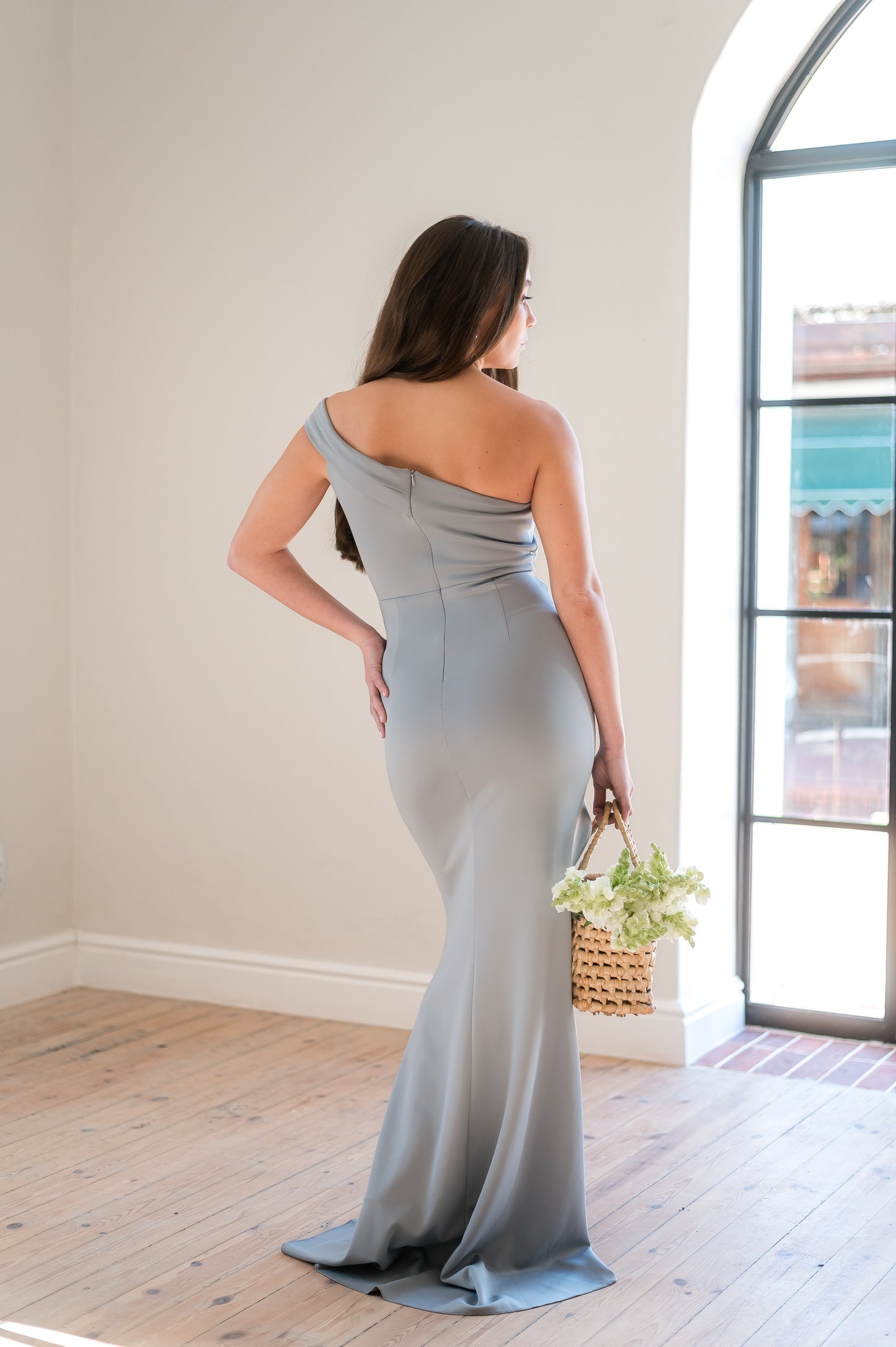 Woman in a sage bridesmaid dress featuring a one - shoulder design in a light filled room.