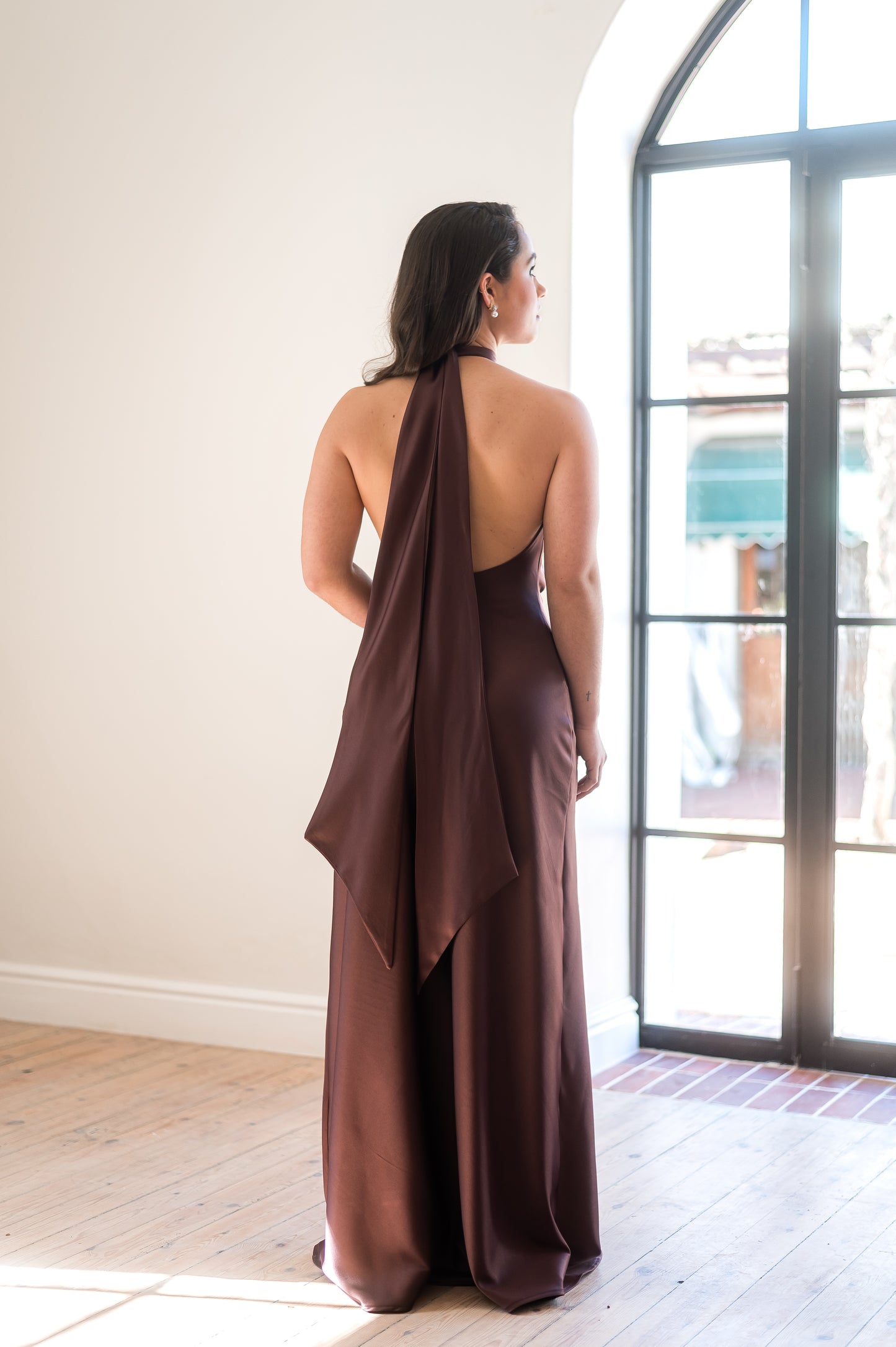 Woman in a coffee brown, open back Bridesmaids dress with dramatic tails standing in a room with a large window.