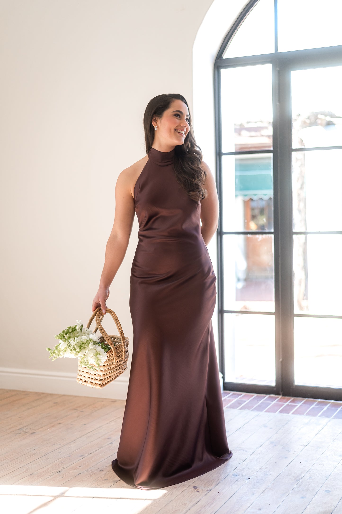 Woman in a coffee brown halter neck bridesmaids dress holding a basket with flowers indoors.