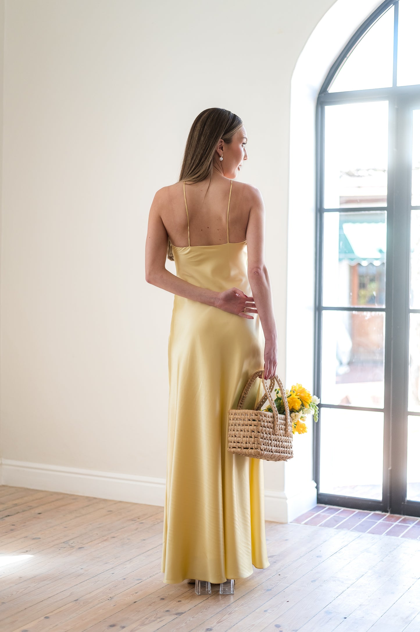 Woman in a butter yellow dress holding a basket with flowers, standing in a room with a large window.
