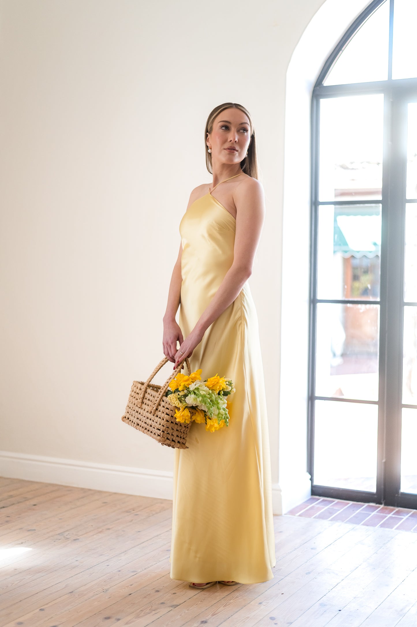 Woman in a butter yellow, halter neck bridesmaid dress holding a basket of flowers indoors.
