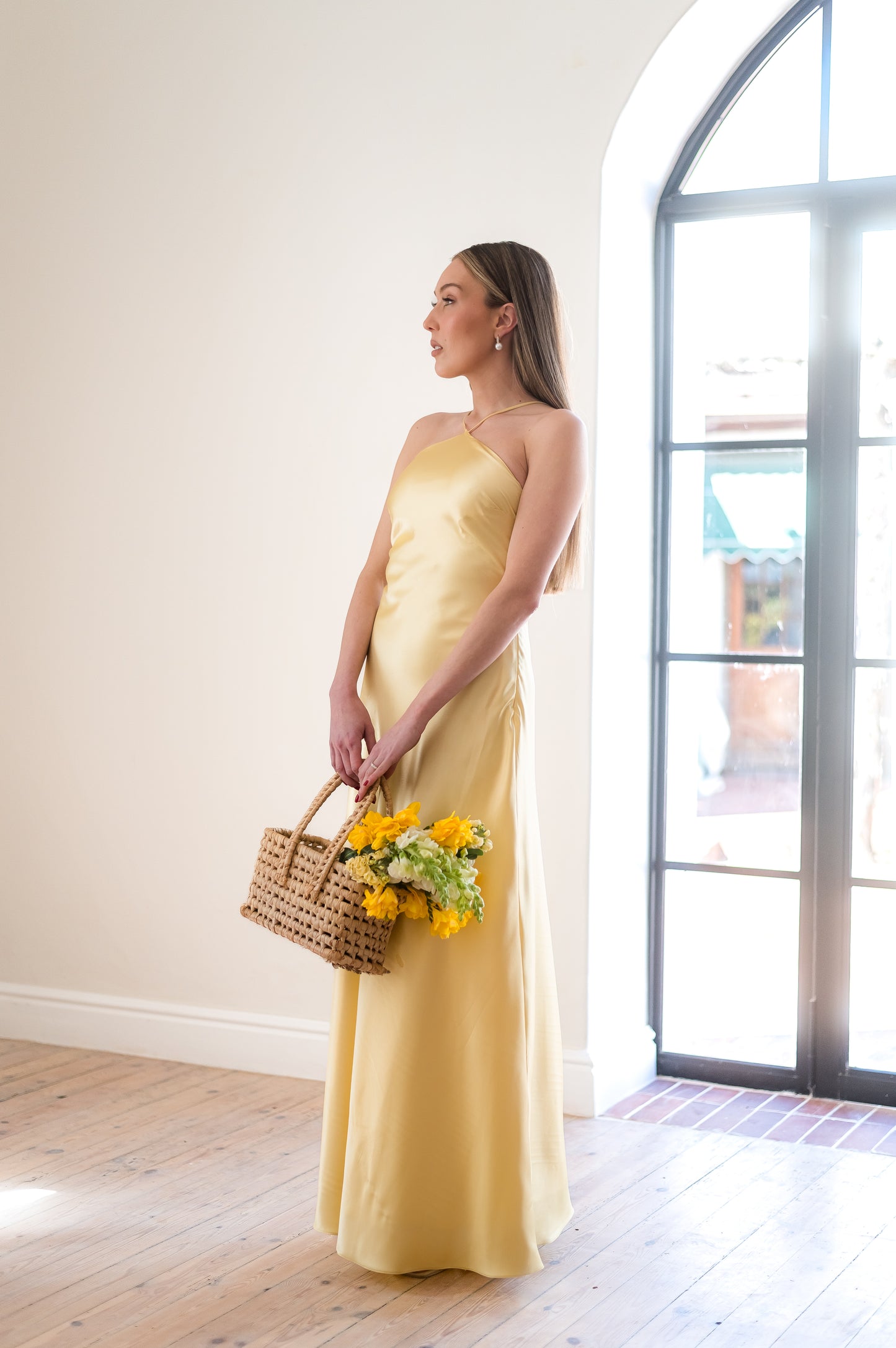 Woman in a butter yellow, satin dress holding a basket of flowers in a light filled room.