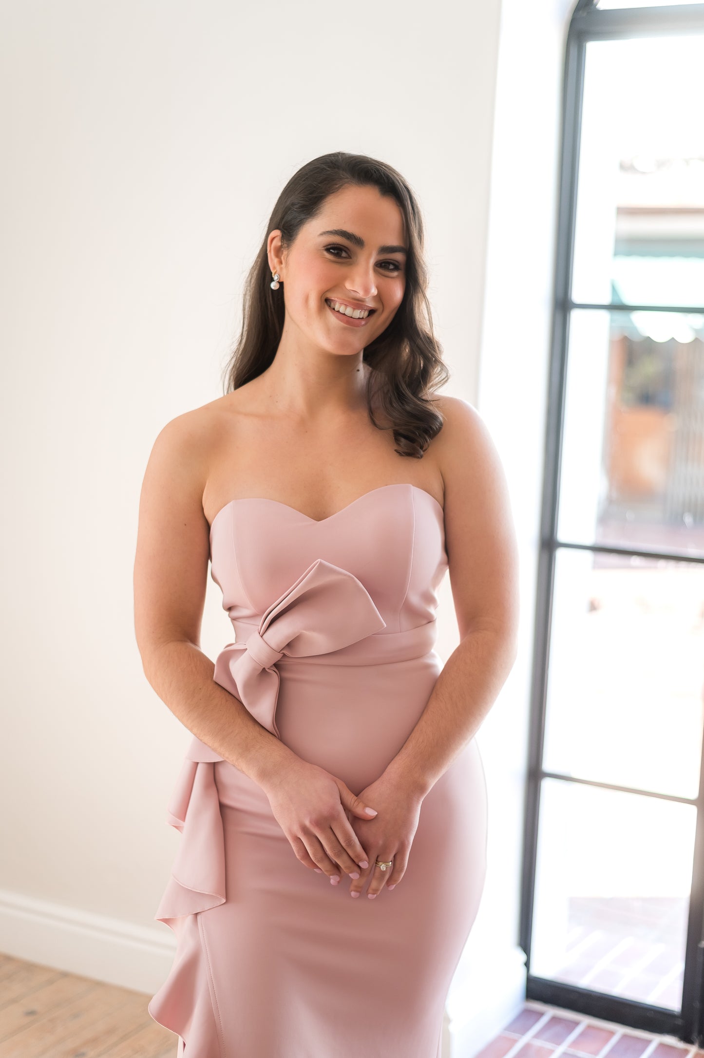 Close up of a woman standing in a dusty pink bridesmaid dress featuring a sweetheart neckline, bow and ruffle detail in cut-out slit.