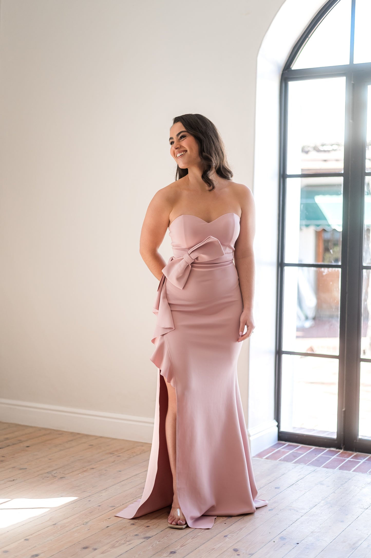 Woman wearing a dusty pink, sweetheart neckline, strapless bridesmaid dress with a bow detail in a room with wooden floor and large window.
