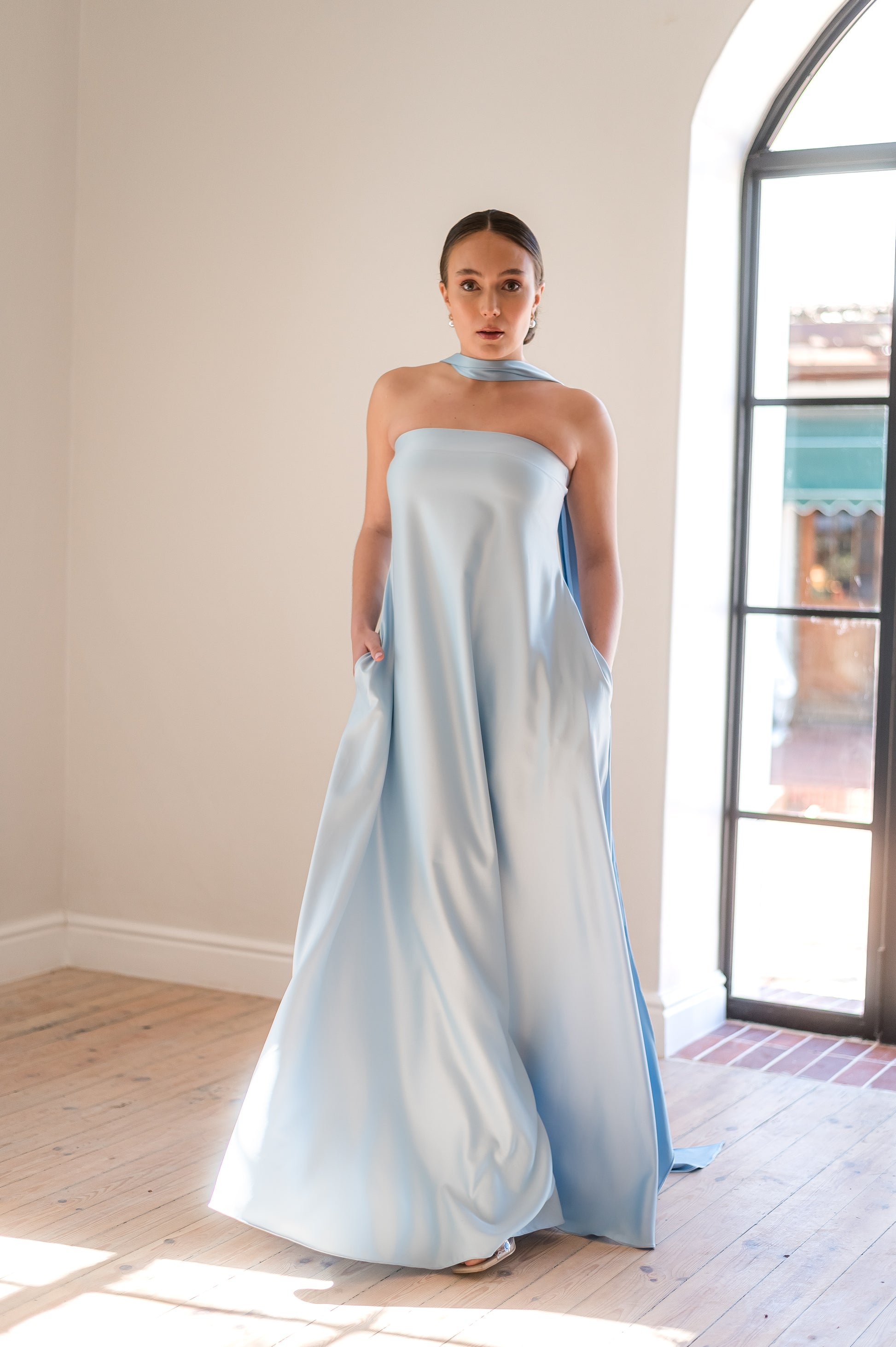 Woman wearing a light blue strapless gown with pockets and neck scarf  standing in a room with wooden flooring and a large window.