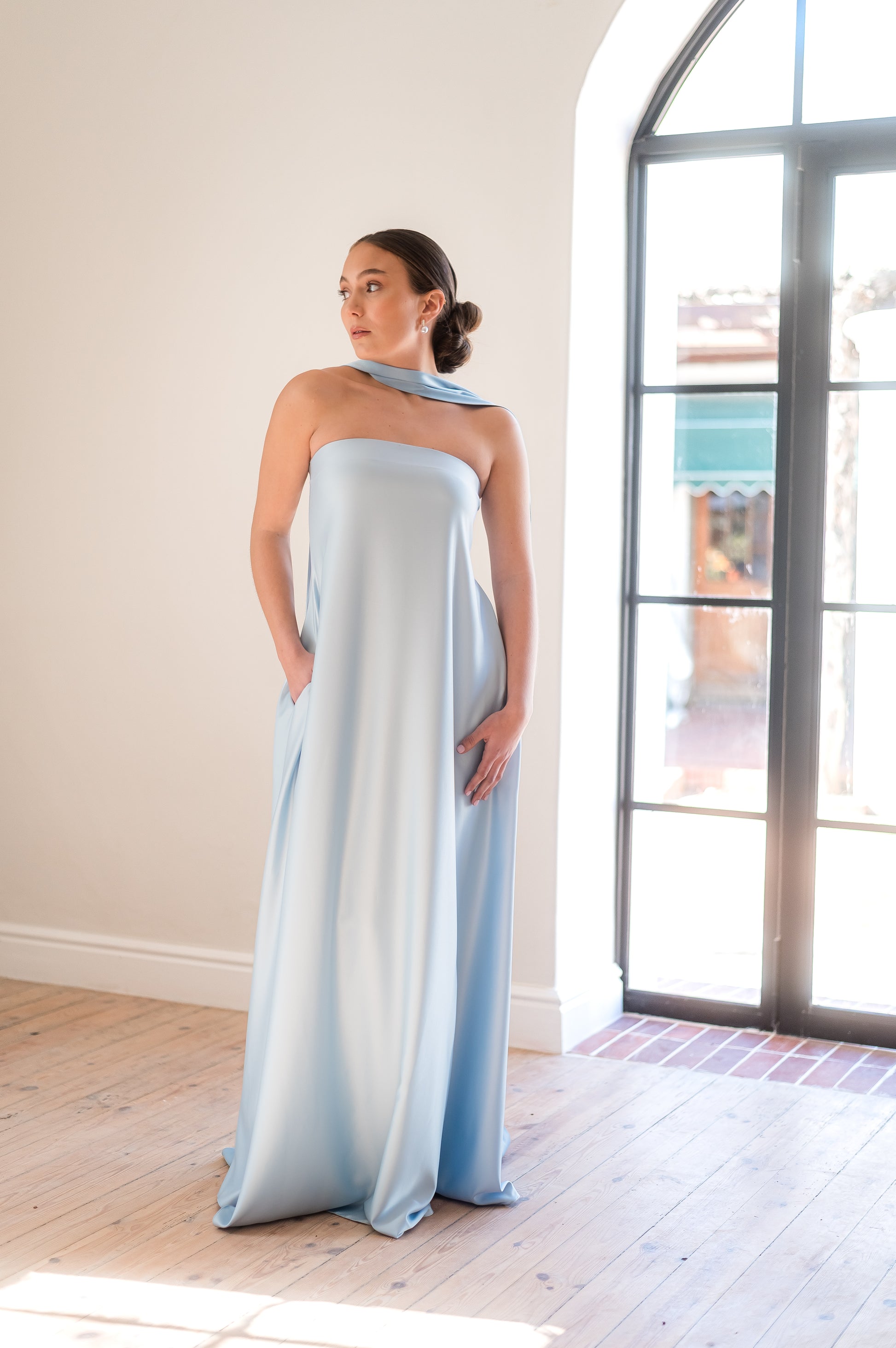 Woman wearing a light blue strapless dress with a neck scarf in a room with large windows.