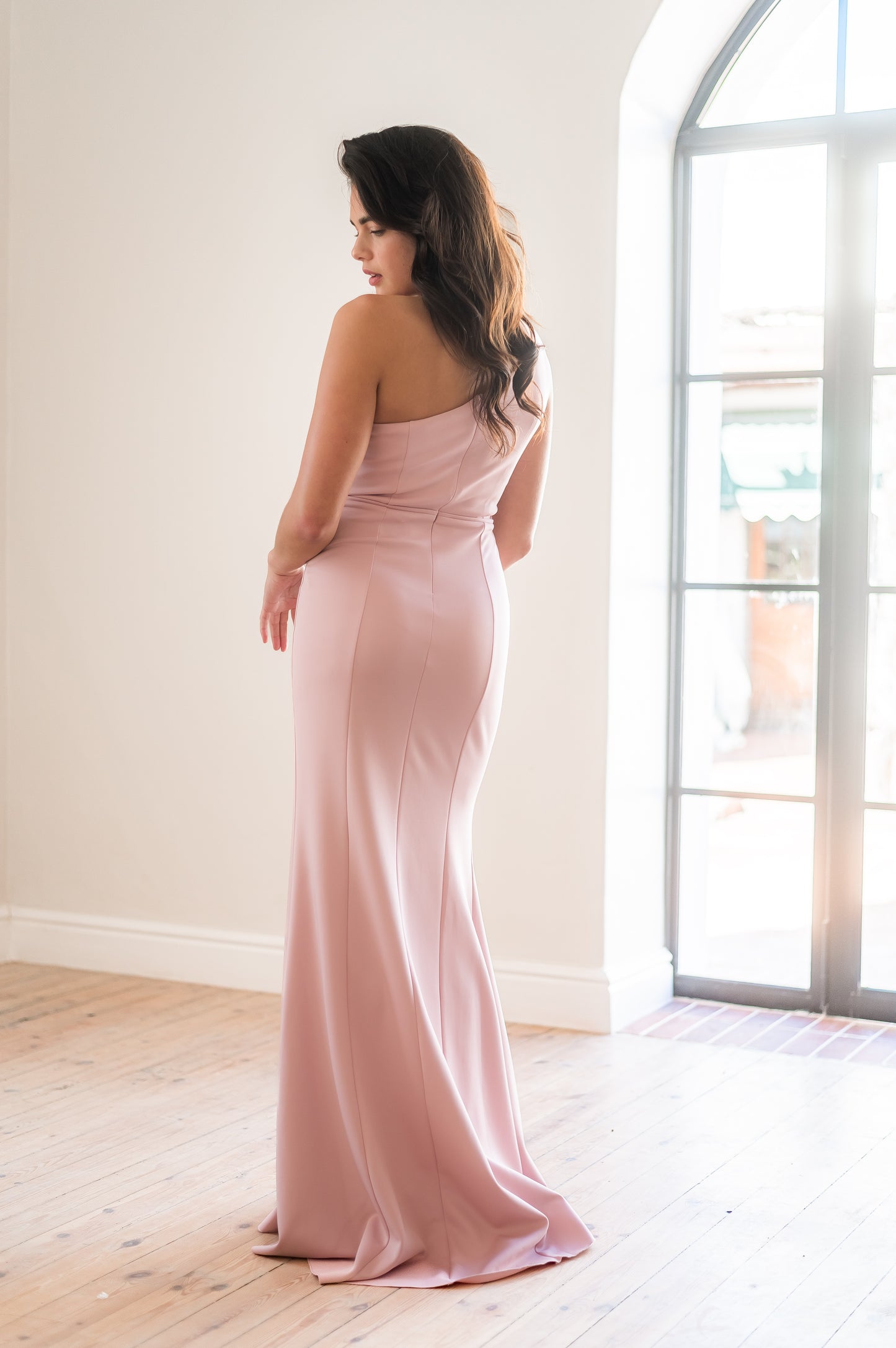 Woman in a pink one-shoulder bridesmaid dress standing in a bright room with large windows.