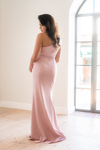 Woman in a pink one-shoulder bridesmaid dress standing in a bright room with large windows.