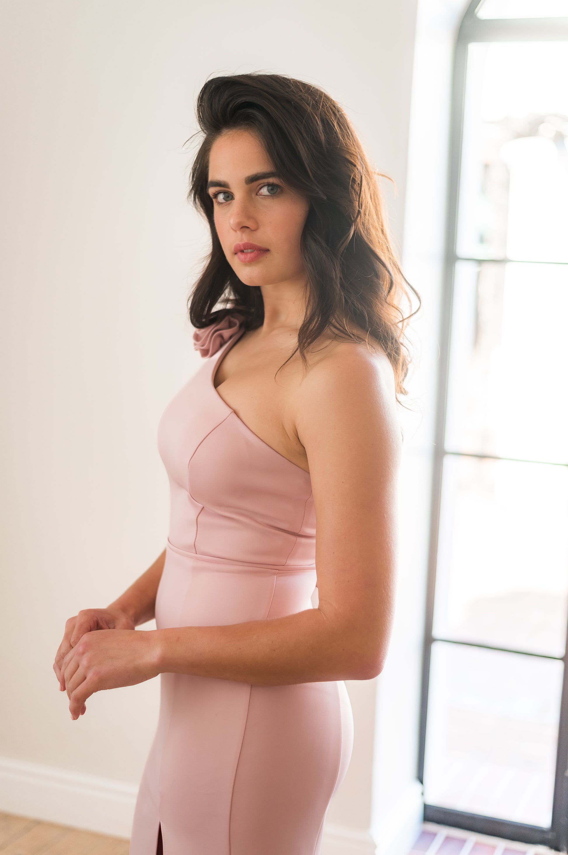 Woman wearing a pink one shoulder bridesmaid dress standing indoors with a neutral background