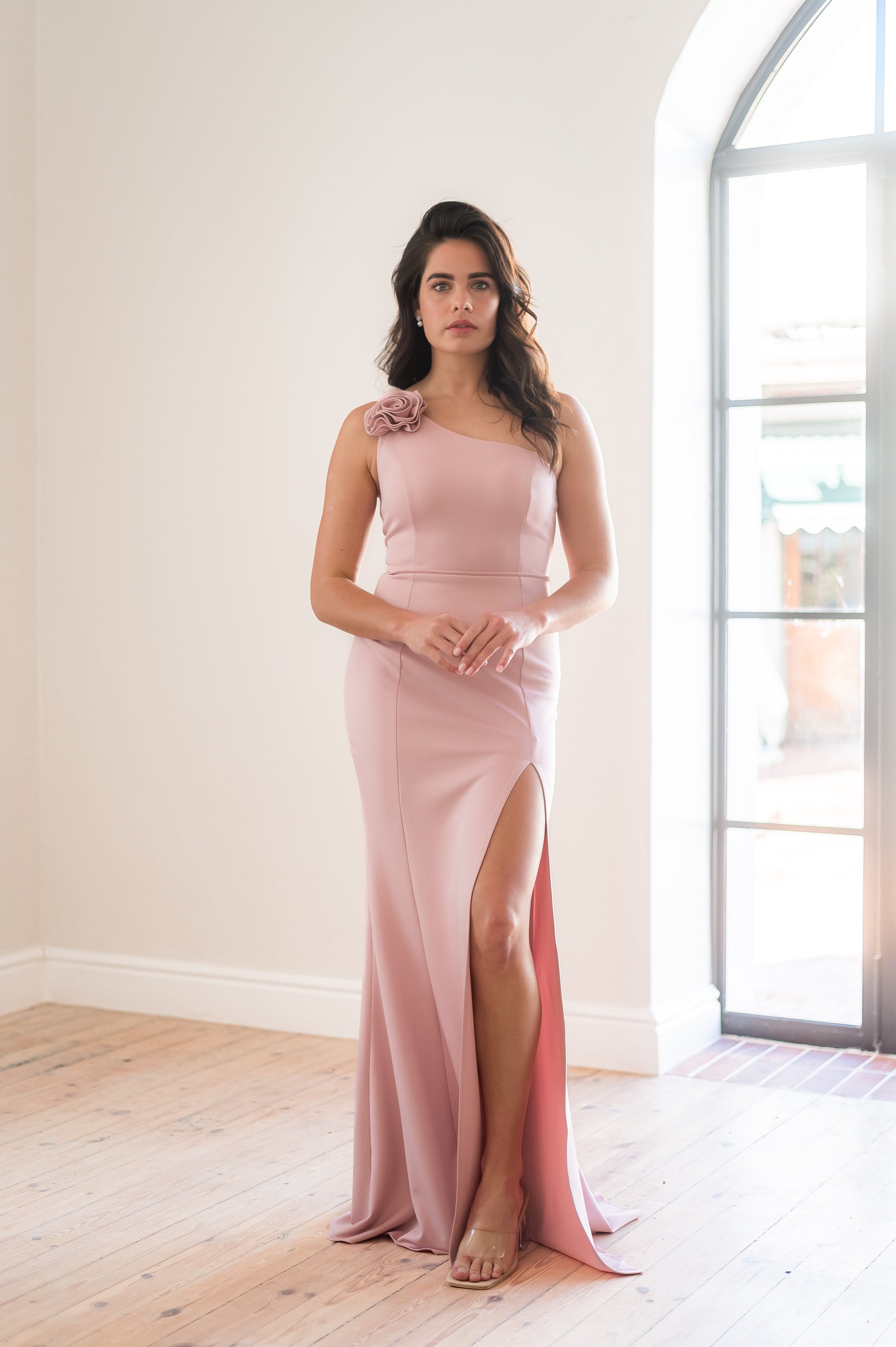 Woman wearing a pink one shoulder bridesmaid dress with a thigh slit standing in a light filled room. 