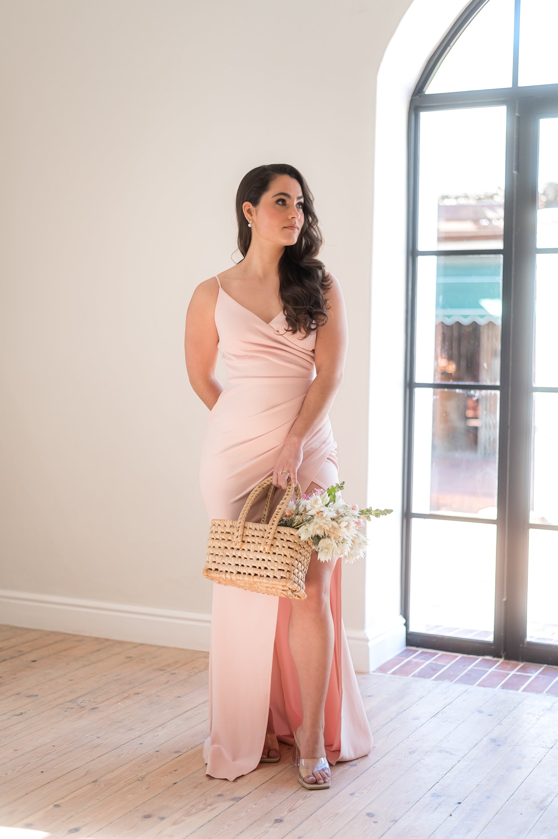 Woman standing in a dusty pink bridesmaid dress featuring thin straps, a V-neck, 
Rouging over tummy and a Cross-over slit.
