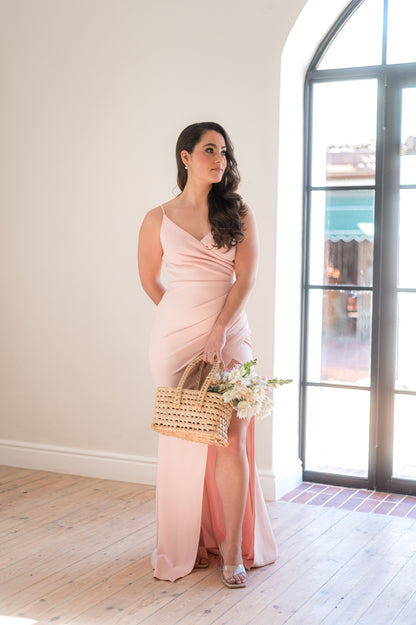 Woman standing in a dusty pink bridesmaid dress featuring thin straps, a V-neck, 
Rouging over tummy and a Cross-over slit.