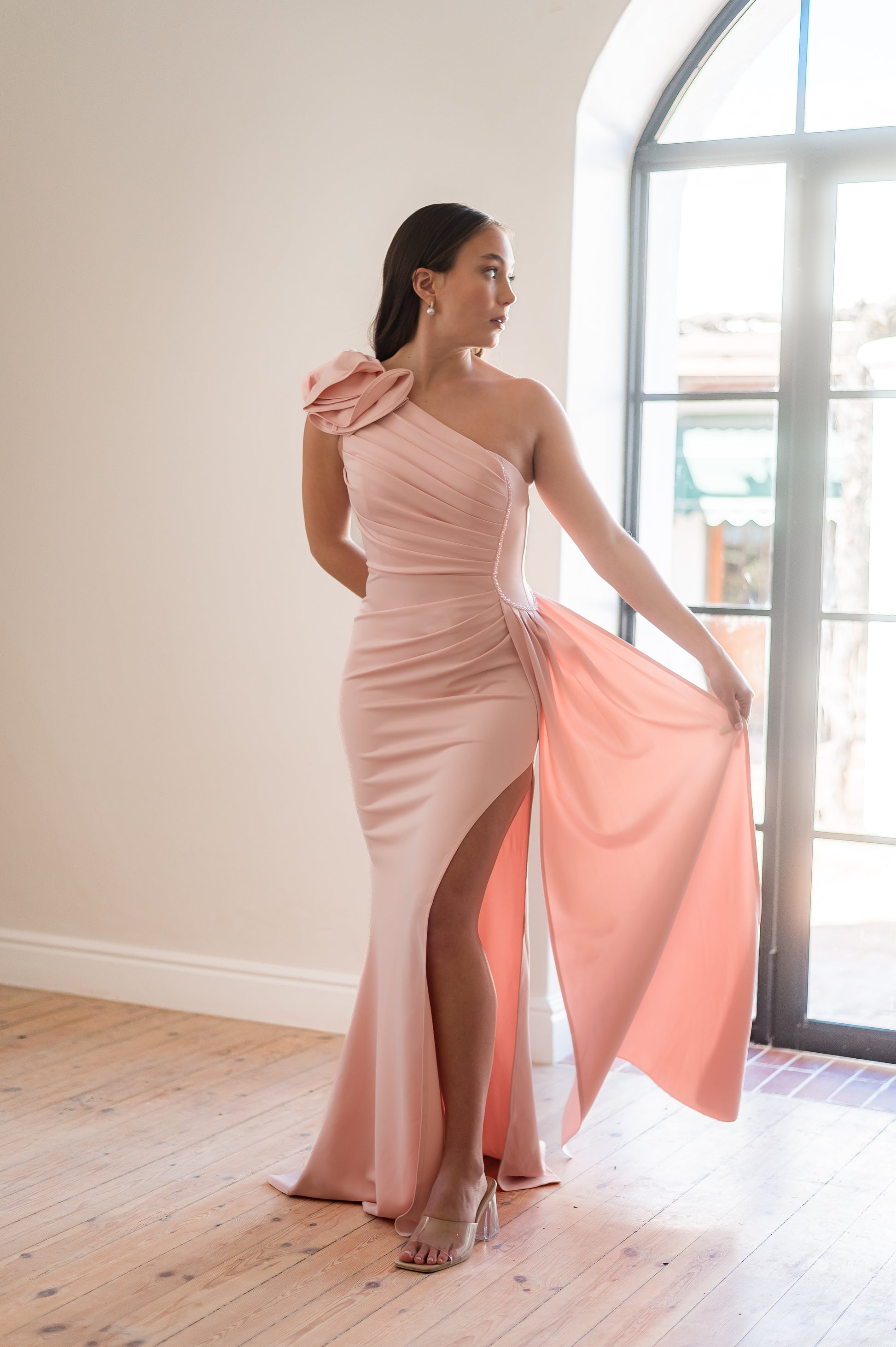 Woman wearing a pink one-shoulder, 3D flower detail  bridesmaids dress with beading on the one side of the waist. 
