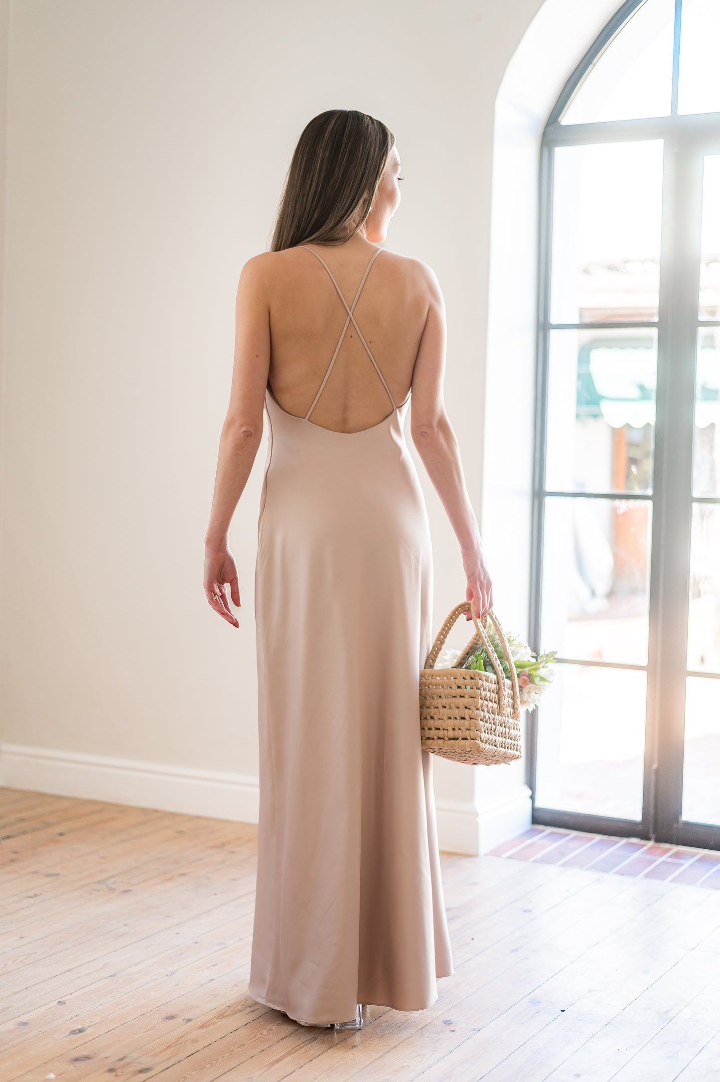 Woman in a long, champagne coloured  bridesmaids dress with a low back and thin straps holding a bouquet indoors.