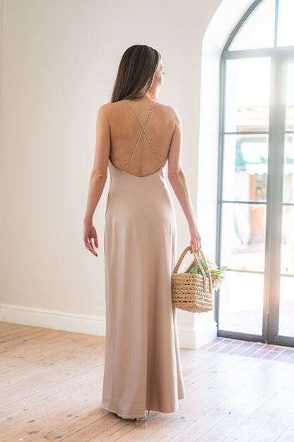 Woman in a long, champagne coloured  bridesmaids dress with a low back and thin straps holding a bouquet indoors.