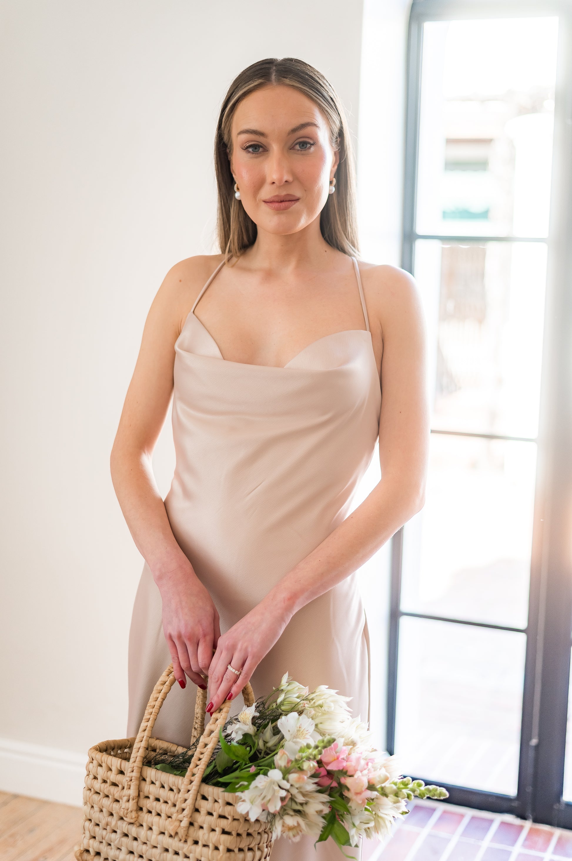 Woman in a satin , champagne coloured  bridesmaids dress with a cowl neckline holding a bouquet indoors.