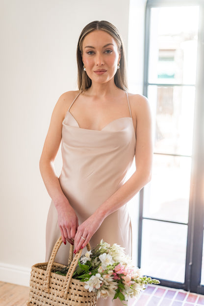 Woman in a satin , champagne coloured  bridesmaids dress with a cowl neckline holding a bouquet indoors.