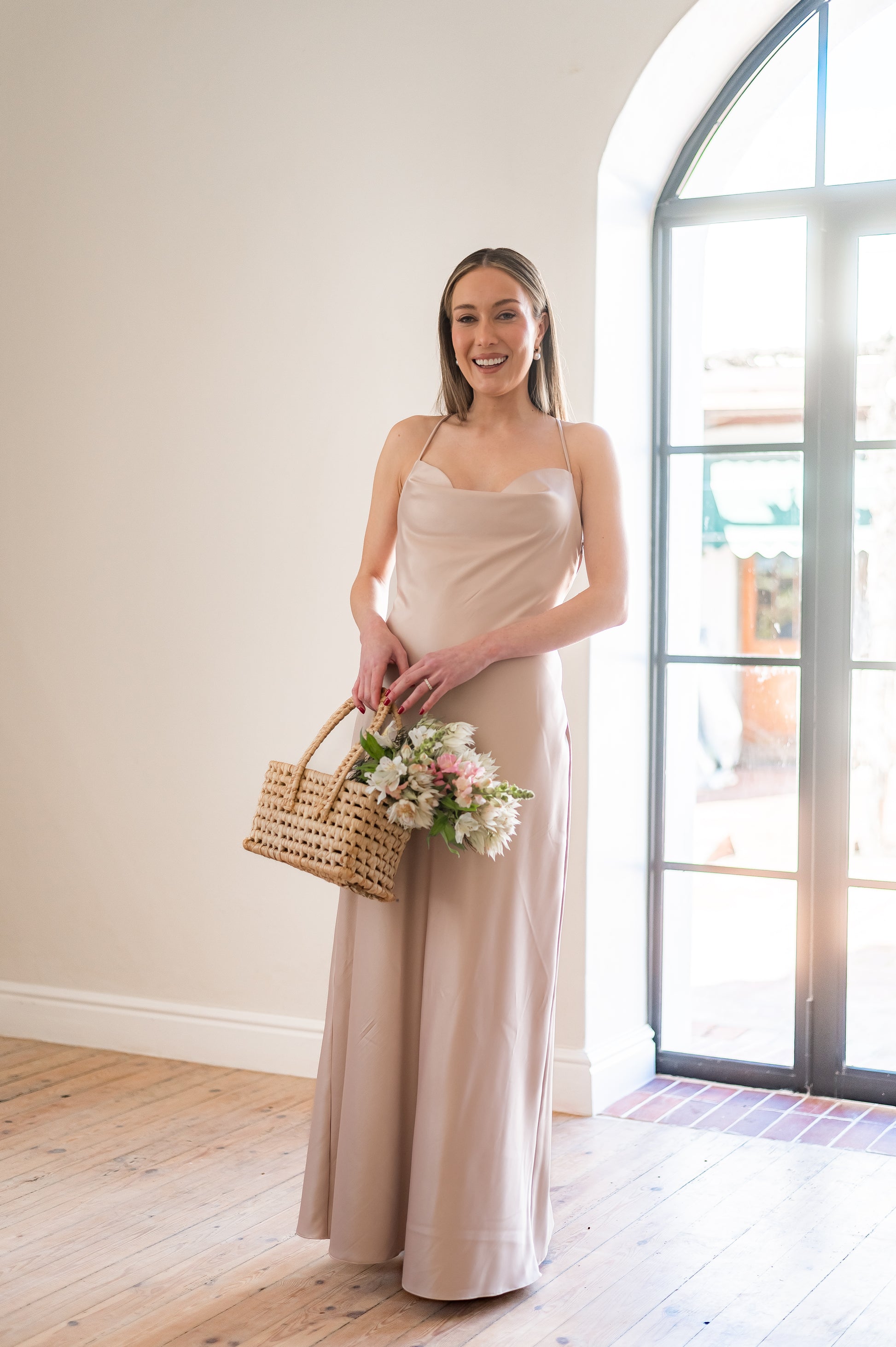 Woman in a long, champagne coloured  bridesmaids dress with a cowl neckline holding a bouquet indoors.