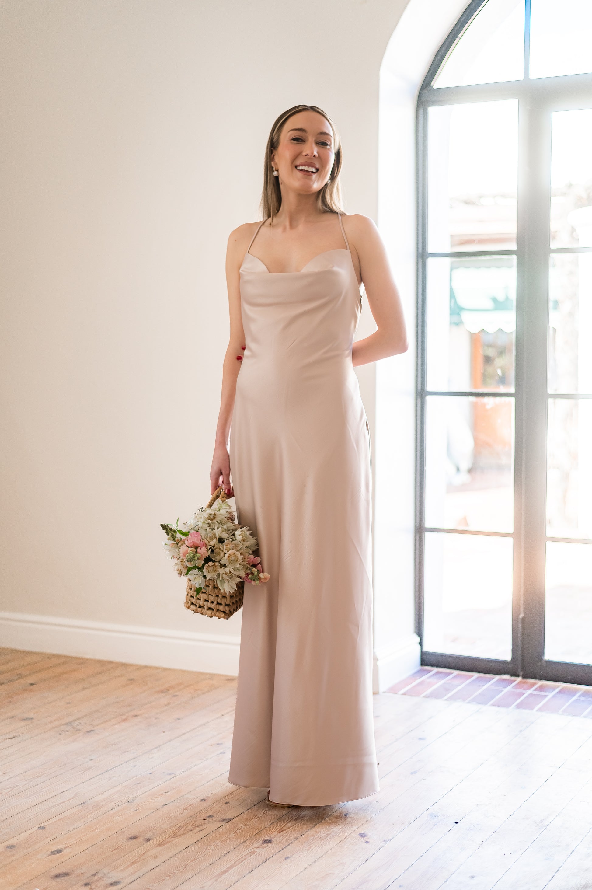 Woman in a long, champagne coloured  bridesmaids dress with a cowl neckline holding a bouquet indoors.