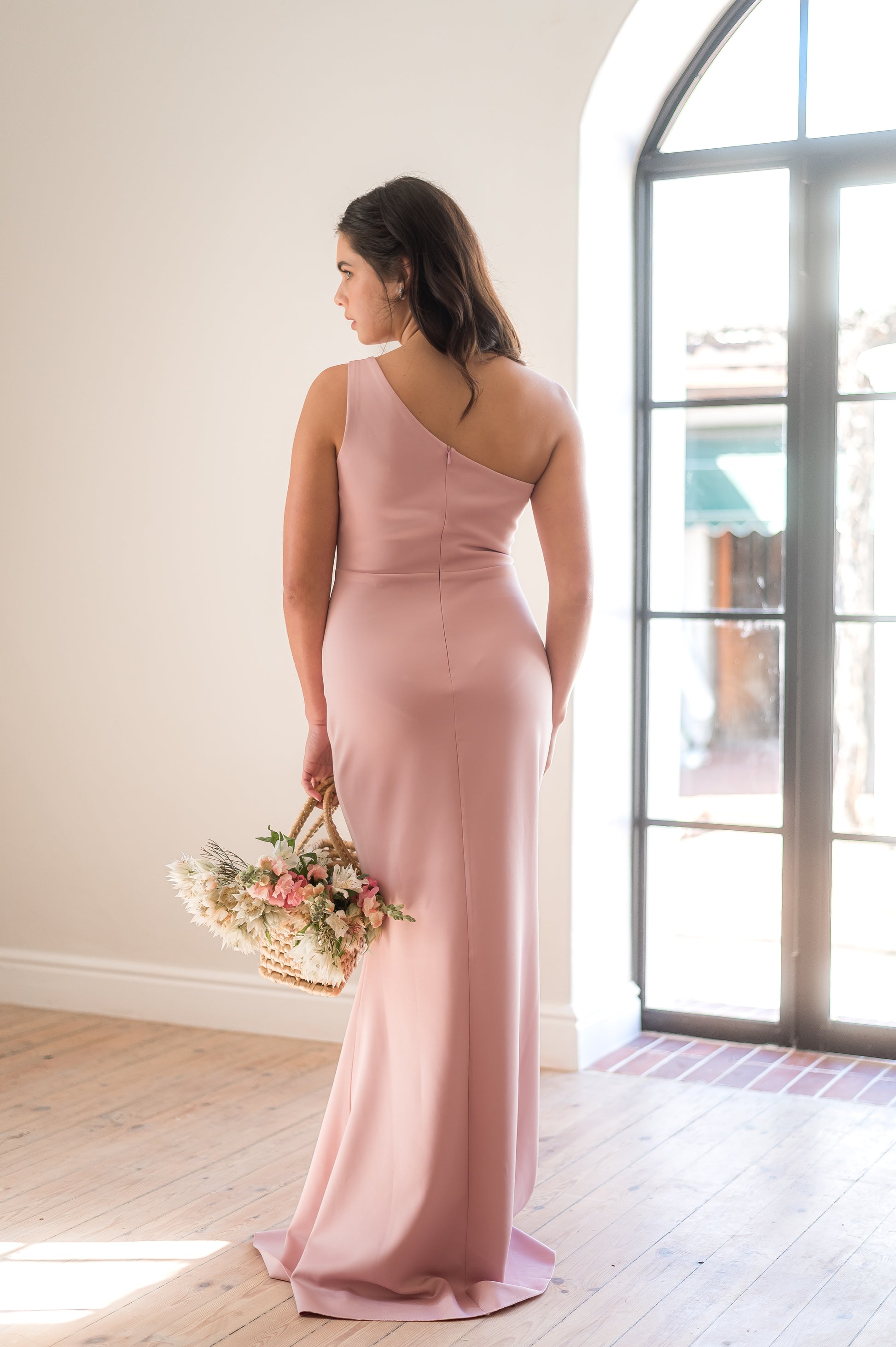Woman standing in a one-shoulder, full length dusty pink bridesmaid dress from the back. 
