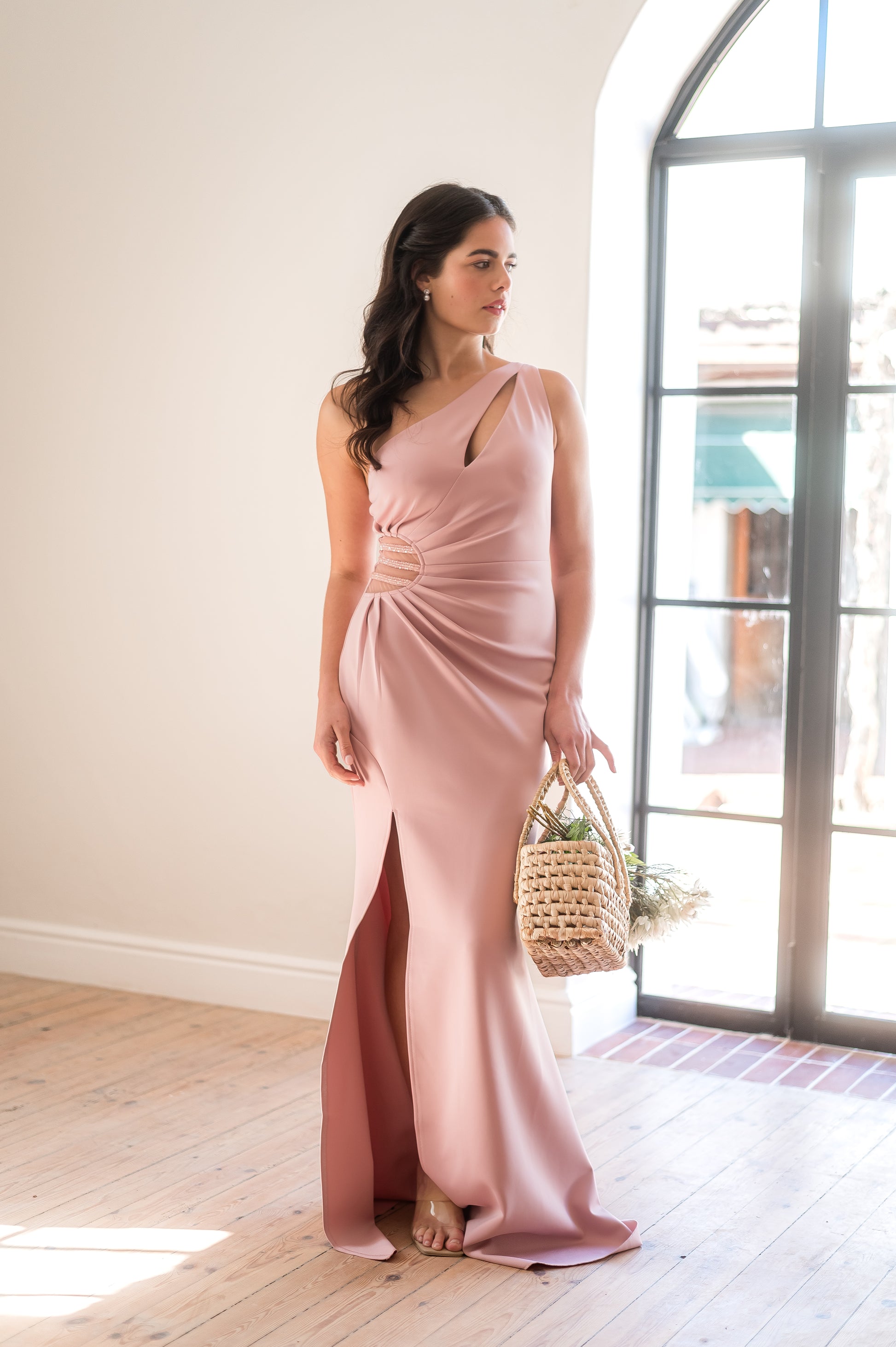 Woman in a dusty pink bridesmaid dress with a one-shoulder neckline, beading in the waist, rouging over the tummy and a thigh slit standing in a room with wooden flooring and large windows.