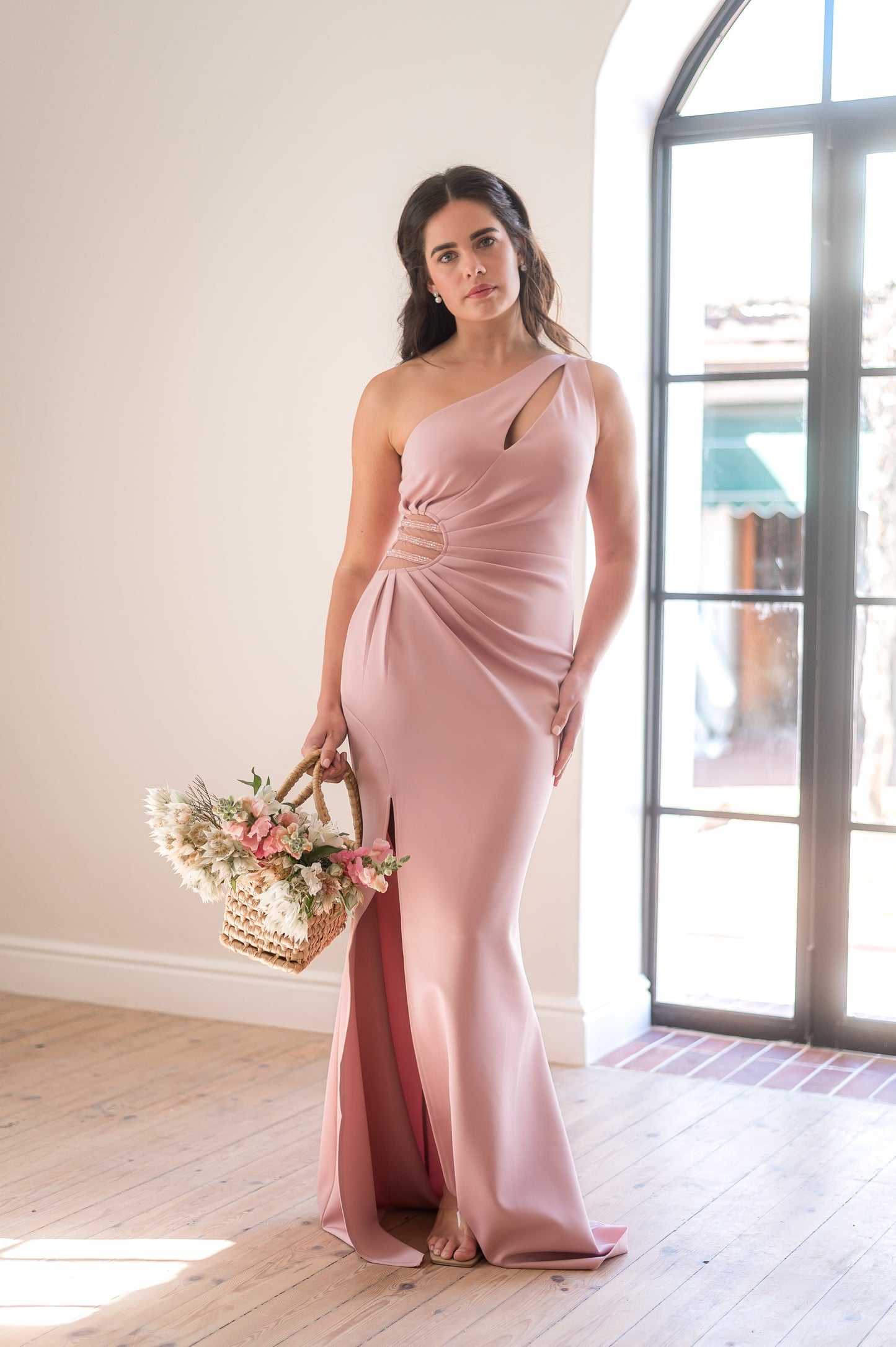 Woman in a dusty pink bridesmaid dress with a one-shoulder neckline, beading in the waist, rouging over the tummy and a thigh slit standing in a room with wooden flooring and large windows.