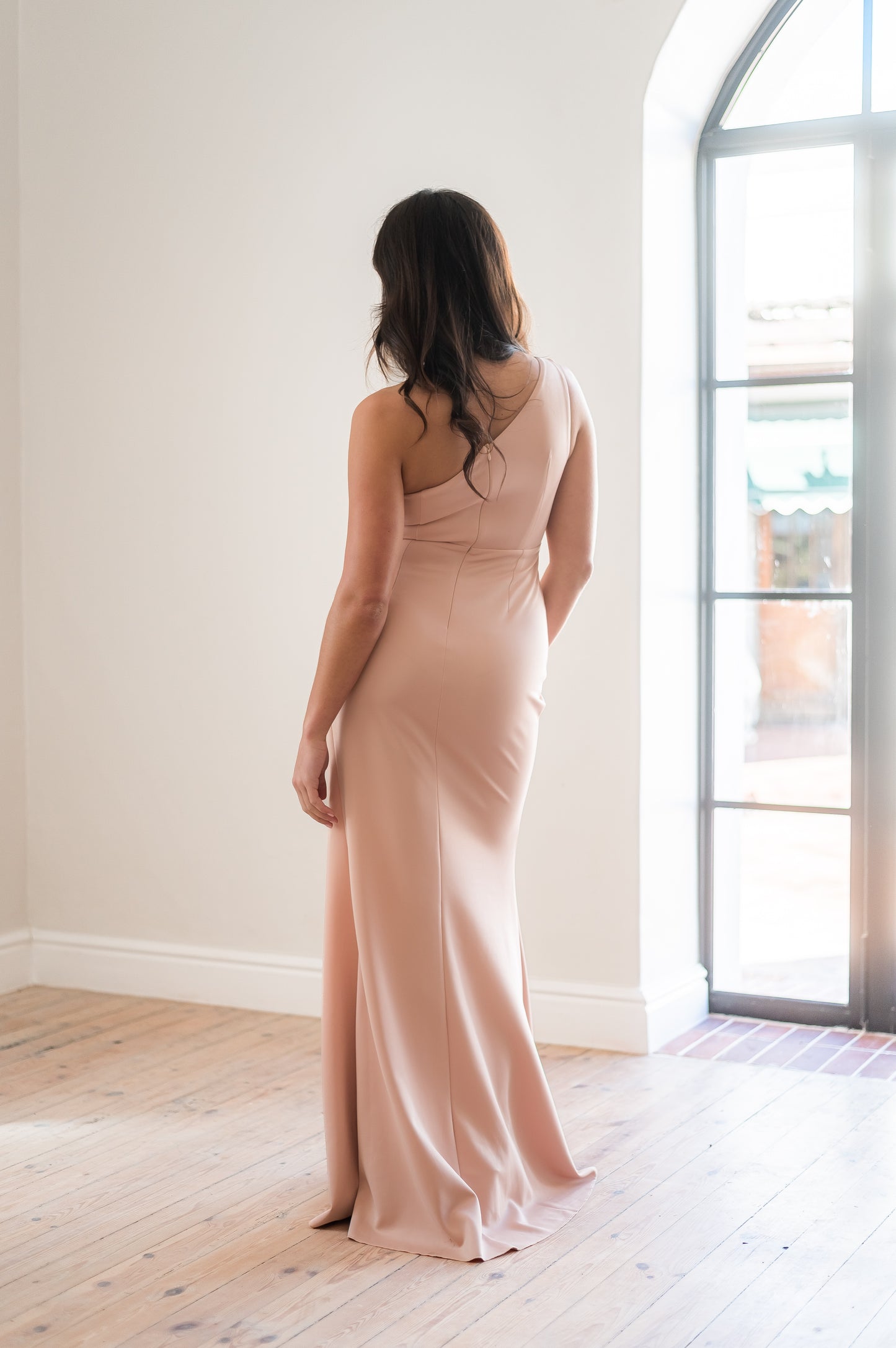 Woman wearing a long dusty pink  one-shoulder bridesmaid dress from the back standing indoors with a neutral background.  