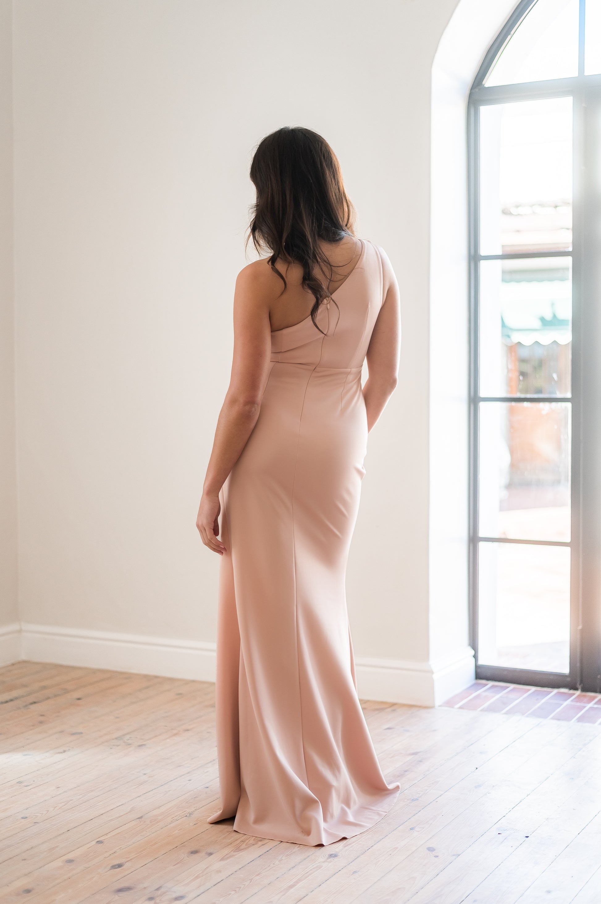 Woman wearing a long dusty pink  one-shoulder bridesmaid dress from the back standing indoors with a neutral background.  