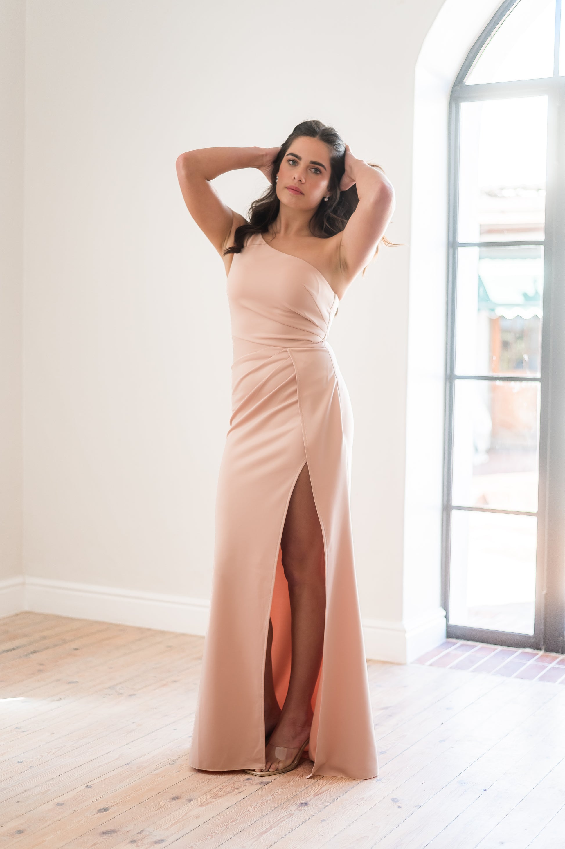Woman wearing a long dusty pink  one-shoulder bridesmaid dress featuring rouging over the tummy and a cross - over slit  indoors with a neutral background. 