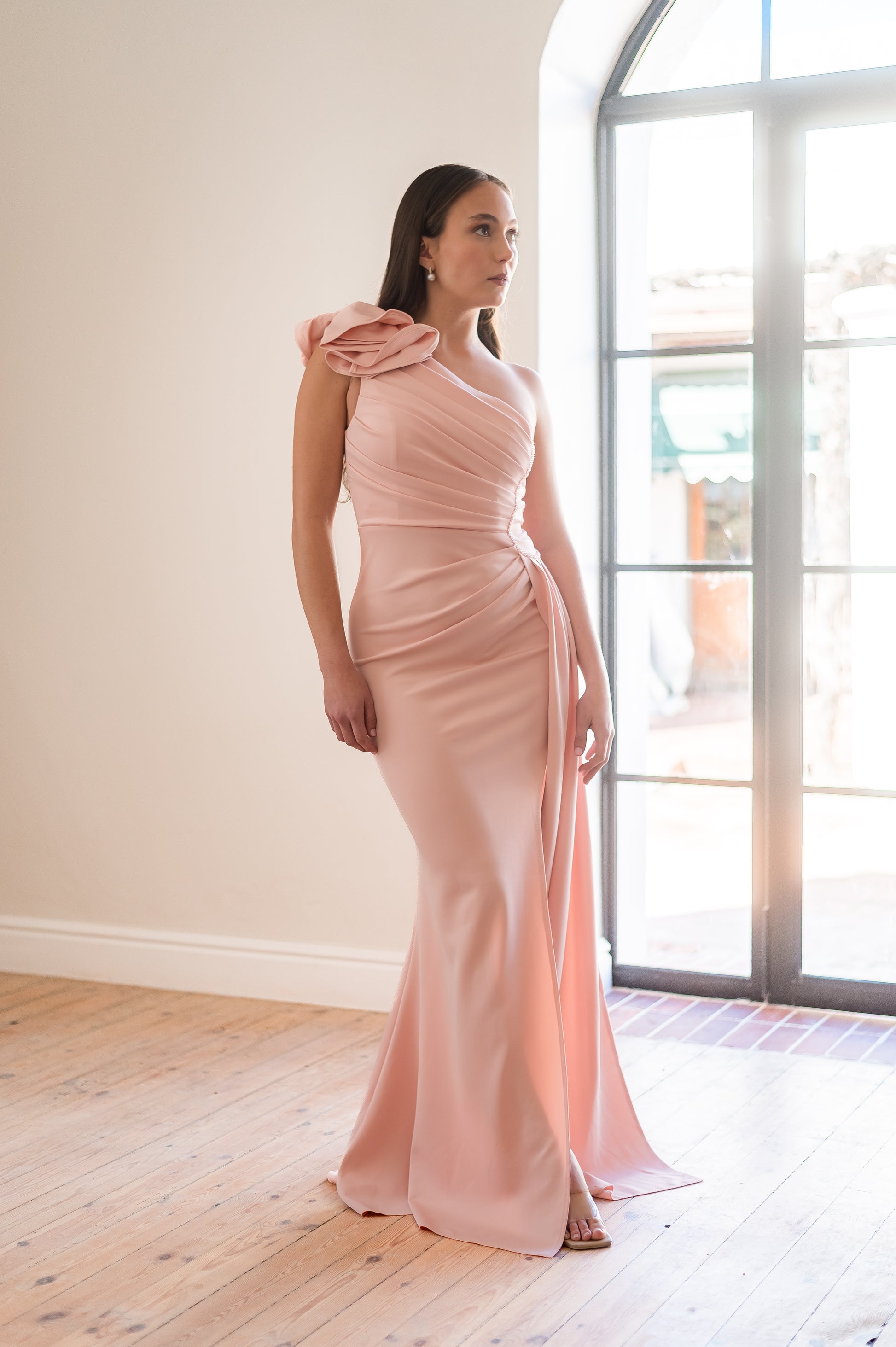 Woman wearing a pink one-shoulder, 3D flower detail  bridesmaids dress with beading on the one side of the waist. 